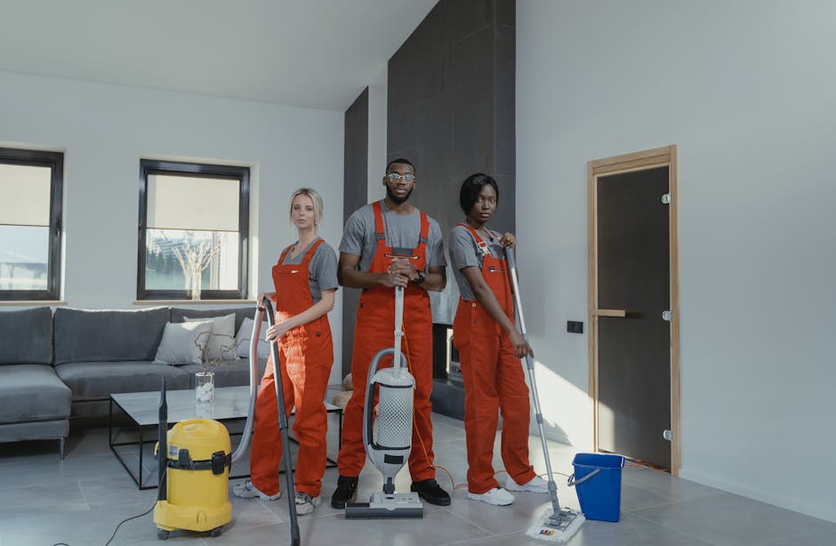 Two professional cleaners from Lambeth Cleaner standing back-to-back against a plain white wall, dressed in beige uniforms. The cleaner on the left, of Asian descent, holds a red vacuum cleaner hose,