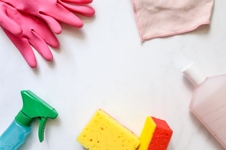 An arrangement of cleaning tools and supplies for surface cleaning and sanitisation in a bright, well-lit setting. There are pink rubber gloves at the top left corner, a pink cloth in the top right corner, and a translucent spray bottle in the bottom left corner. A pink and yellow sponge are positioned at the bottom center, with a white bottle of cleaning liquid on the right side. The surfaces and materials include smooth, shiny plastic, fabric, and sponge textures, indicating a focus on domestic cleaning. The overall cleanliness appears maintained, with no visible dust or dirt, reflecting professional deep cleaning practices. Lambeth Cleaner emphasizes thorough sanitisation for residential and commercial spaces, aligning with their focus on effective hygiene and surface care.