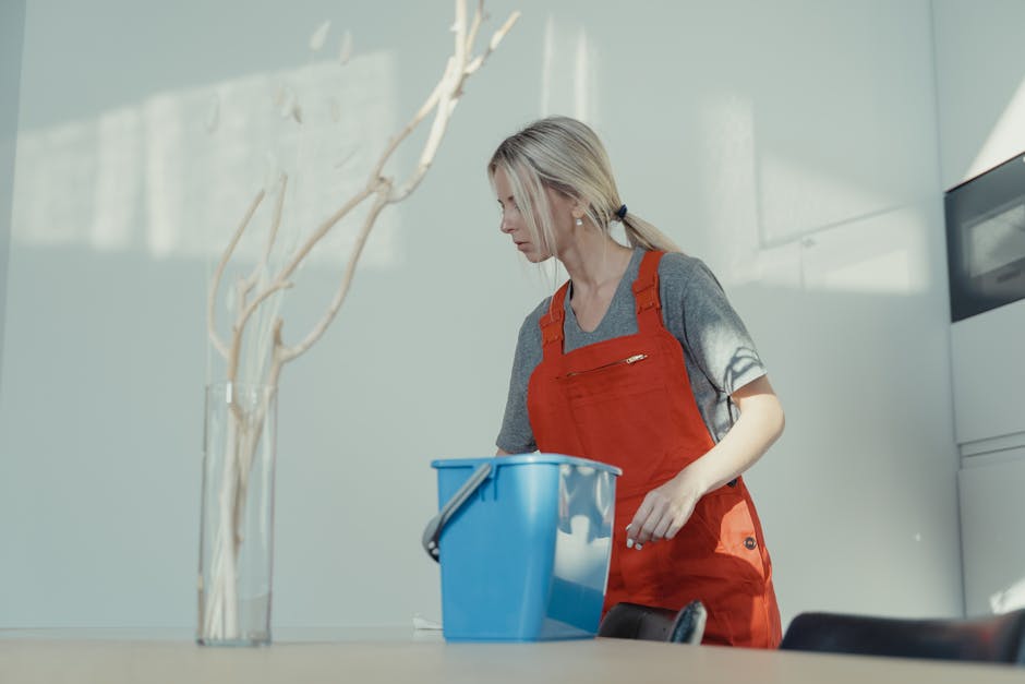 A woman with blonde hair tied back, dressed in a grey T-shirt and red cleaning apron, is engaged in surface cleaning inside a bright, modern room. She is leaning slightly forward, focusing on the task at hand, with her right hand resting on a blue plastic bucket. Visible surfaces include a smooth, light-colored countertop and a white wall in the background, illuminated by natural daylight. There are minimal visible tools or cleaning products, emphasizing a neat and hygienic environment. The room features a sparse, contemporary design, with a decorative plant in the foreground contributing to the aesthetic. The clean, shiny surfaces and well-lit setting reflect a focus on effective domestic cleaning, aligned with the insider tips for landlords on the Brixton flat cleaning page by Lambeth Cleaner.