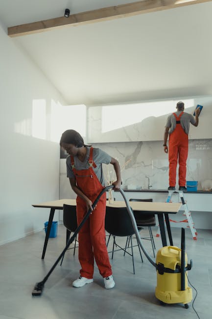 Two professional cleaners from Lambeth Cleaner are performing deep cleaning in a modern residential kitchen. The female cleaner in the foreground is using a vacuum cleaner on a tiled floor, which appe