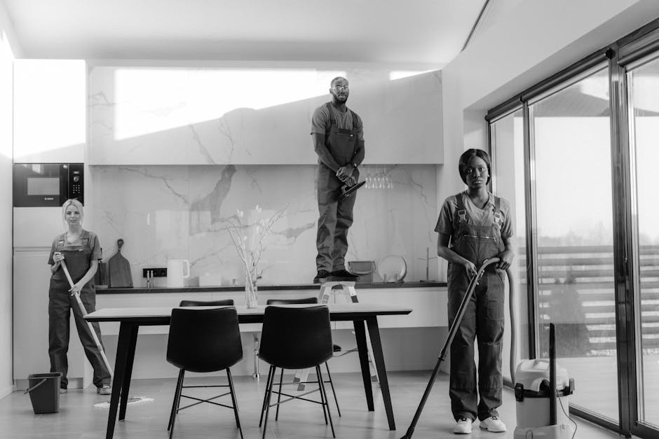 Two professional cleaners from Lambeth Cleaner are performing deep cleaning in a modern residential kitchen. The female cleaner in the foreground is using a vacuum cleaner on a tiled floor, which appe